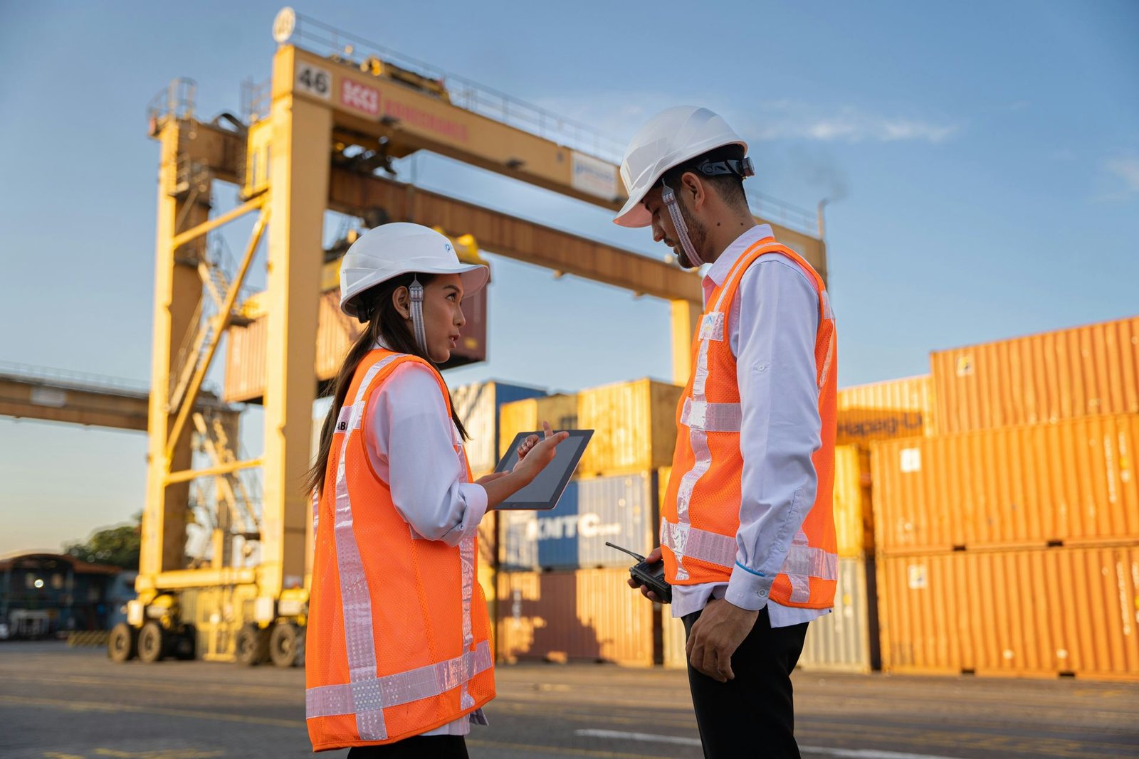 Two logistics coordinators in hi-vis vests reviewing a tablet near shipping containers